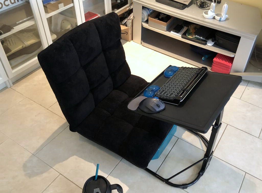 A legless chair sits on the floor, a raised keyboard tray immediately in front of it, and a large mug and straw off to one side.